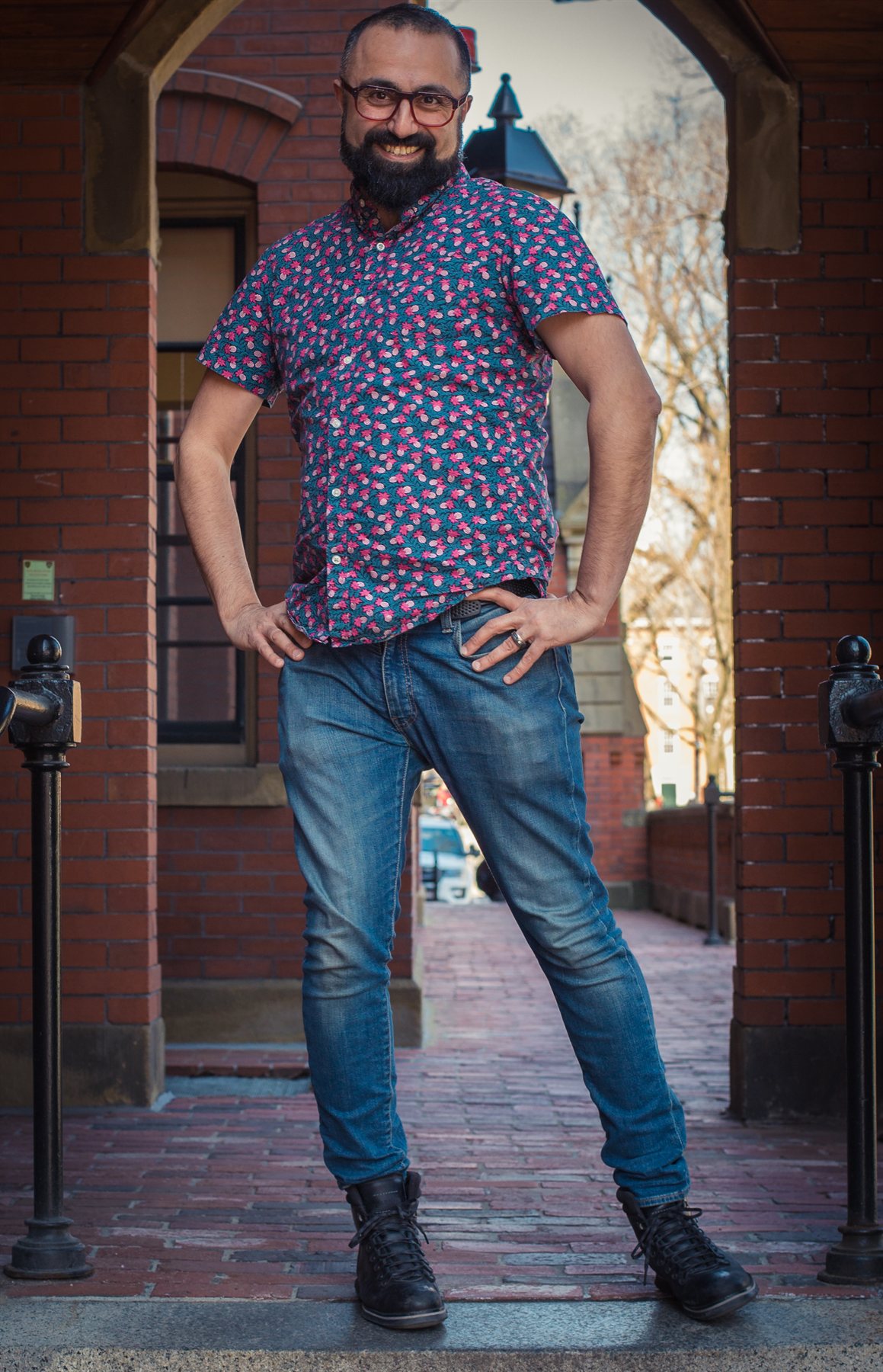 Andy in a blue floral shirt and jeans poses with his hands on his hips beneath a brick archway.