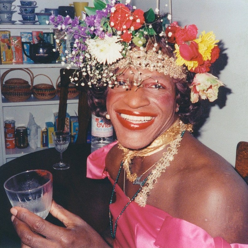 An iconic photo portrait of Marsha Pay-it-no-mind Johnson, a legendary Black queen from the time of Stonewall. She's wearing a satin pink dress that reveals her milk chocolate shoulders. She wears a beautiful crown of flowers, with some sort of gold ribbon or garland around her forehead and also as a choker necklace around her neck. Her makeup is bold but restrained--pink blush & eye shadow, with bright red lipstick. She smiles a wide smile, seemingly overflowing with joy. A bottle of evian water sits on the table behind her, and she sips out of a nick and nora glass looking every bit of the regal queen she is.